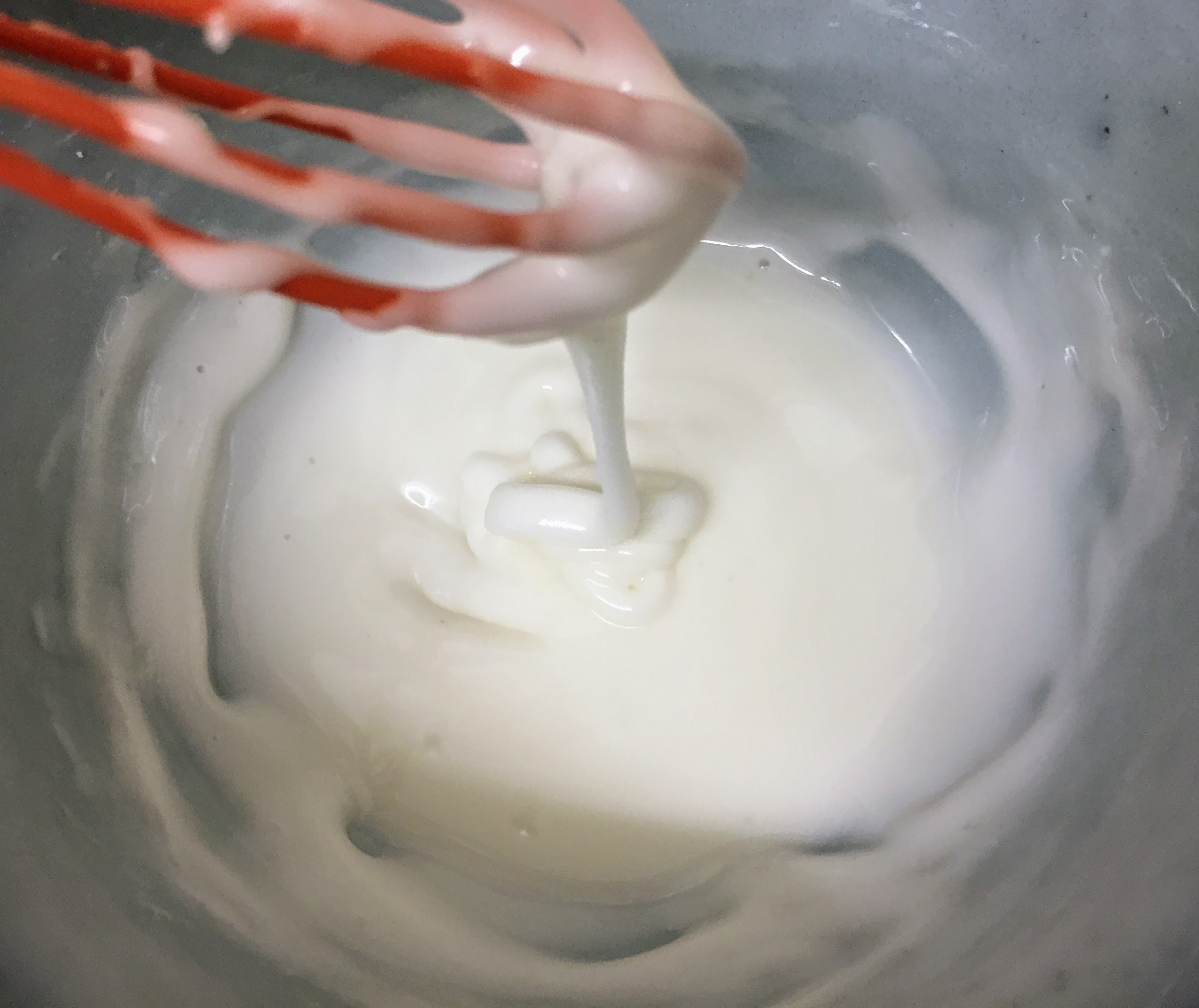 A close up of a bowl of thick white icing. A whisk is held above the bowl so the icing drizzles into the bowl. You can see the trail of the drizzled icing holding it's shape for a few seconds before merging into the rest of the icing, which is the perfect consistency for piping.