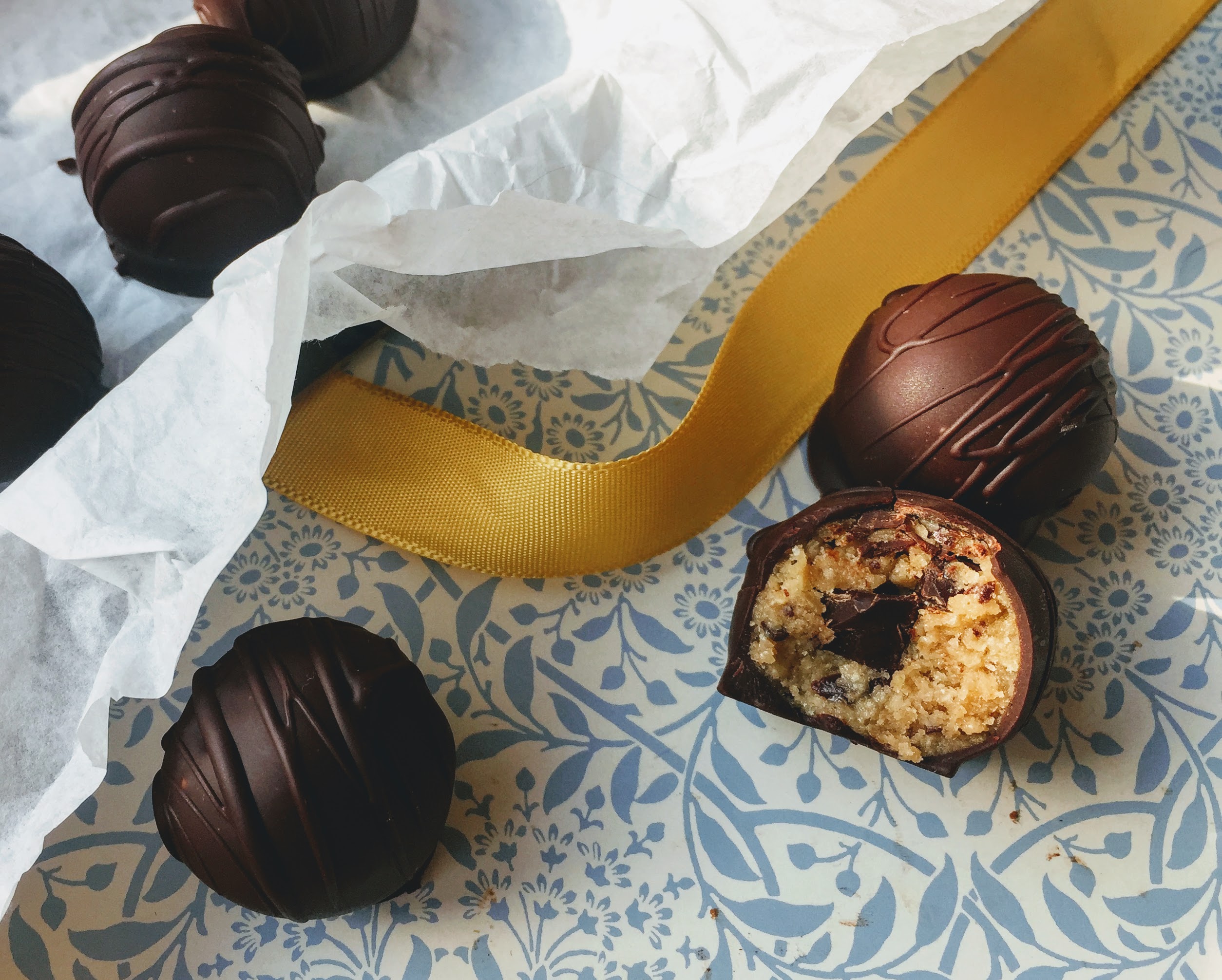 Chocolate-coated cookie dough bites are scattered on a blue floral tray. The chocolate coating is shiny and smooth with streaks of chocolate drizzled on top. One of the chocolates has a bite taken out of it to reveal a golden cookie dough center with small chunks of dark chocolate.